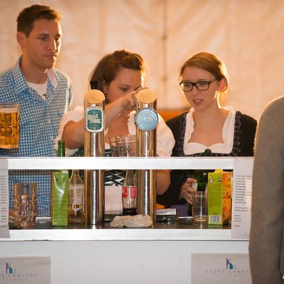 Eine Gruppe von Menschen steht an einer Bar, wo zwei Frauen Getränke servieren und ein Mann ein Glas Bier in der Hand hält. Der Tresen hat Flaschen, Gläser und eine Speisekarte.