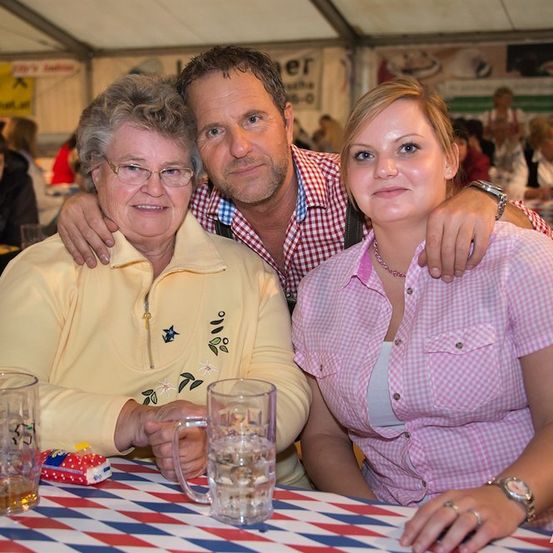 Drei Personen sitzen an einem Tisch mit rot-weiß-blauem Tischtuch. Die Frau links trägt eine Brille und ein gelbes Sweatshirt, während der Mann in der Mitte ein kariertes Hemd trägt. Die Frau rechts trägt ein rosa kariertes Hemd und eine Halskette. Sie lächeln und posieren für ein Foto.