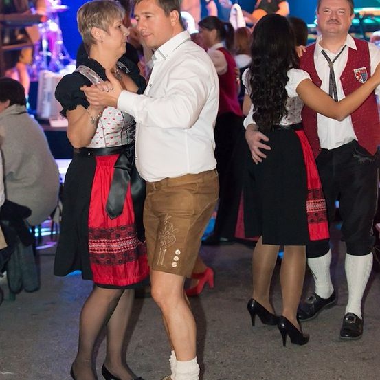 Ein Mann und eine Frau tanzen bei einer lebhaften Veranstaltung zusammen, mit einer Band im Hintergrund. Die Frau trägt ein traditionelles Kleid und hohe Schuhe.
