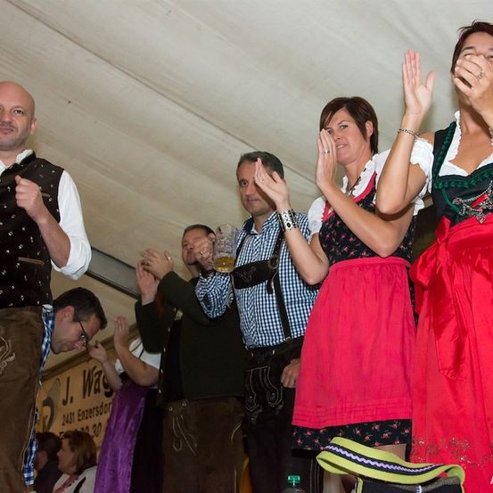 Eine Gruppe von Menschen in traditioneller bayerischer Kleidung klatscht und jubelt bei einer Veranstaltung. Der Mann vorne trägt eine Weste mit Sternmuster.