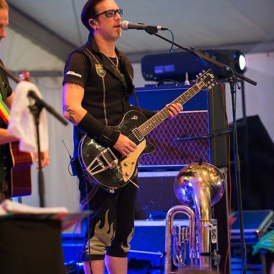 Ein Mann spielt Gitarre auf der Bühne mit einem Mikrofon vor ihm. Eine weitere Person steht hinter ihm mit einer Gitarre. Eine Tuba steht vor dem Lautsprecher.