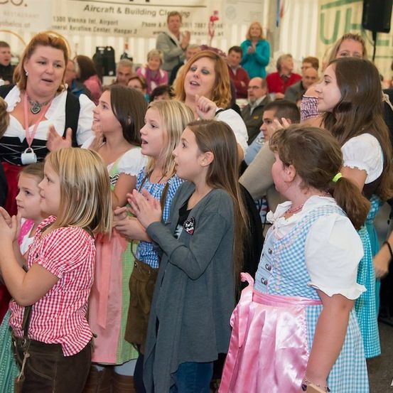 Eine Gruppe von Kindern und Erwachsenen in traditioneller deutscher Kleidung klatscht und singt in einem großen Saal.