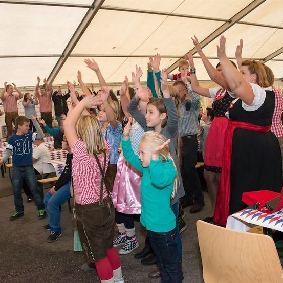 Eine Gruppe von Menschen, einschließlich Kindern, versammelt sich in einem Zelt, hebt die Hände und lächelt. Einige sind in traditioneller Kleidung gekleidet.