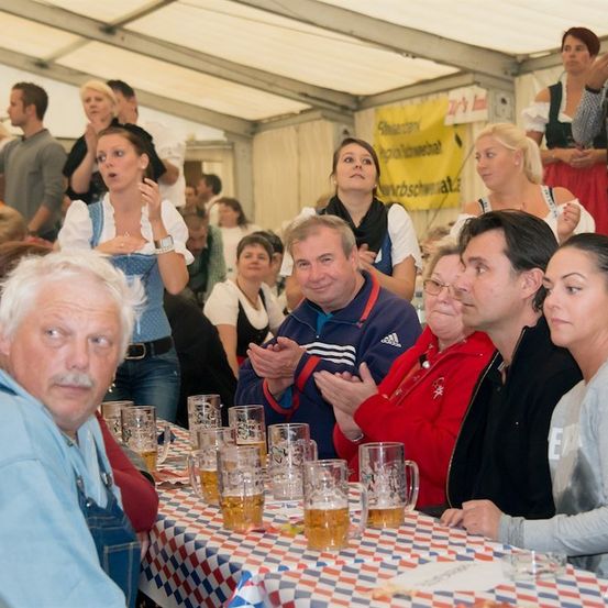 Eine Gruppe von Menschen, einige in traditioneller deutscher Kleidung, versammelt sich um einen Tisch mit Bierkrügen, möglicherweise auf einem Fest.