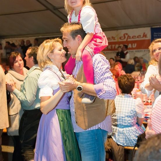 Ein Mann und eine Frau tanzen mit einem kleinen Mädchen auf seinen Schultern bei einem lebhaften Festival. Die Menge ist fröhlich und die Atmosphäre ist festlich.
