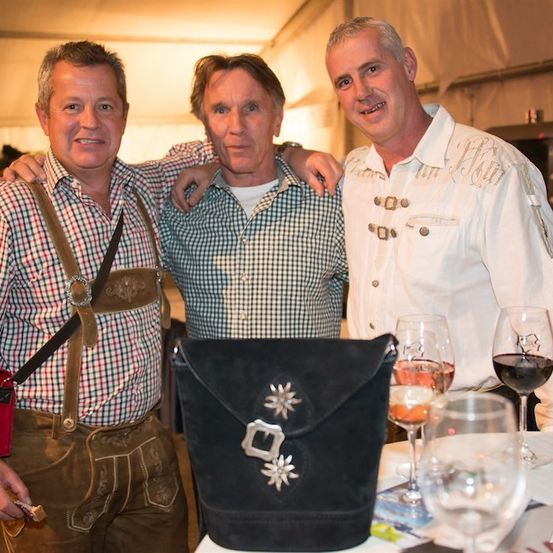 Drei Männer in traditioneller deutscher Kleidung, einer in einem karierten Hemd, ein anderer mit einem weißen Hemd und Hosenträgern und der dritte in einem weißen Hemd mit einer Tasche auf dem Tisch vor ihnen.