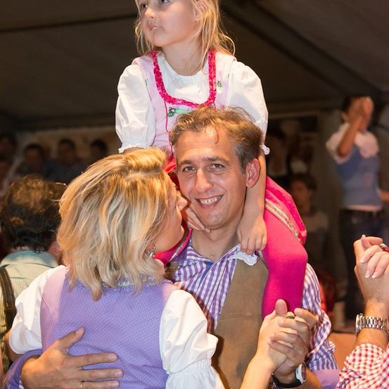 Ein Mann und eine Frau tanzen mit einem kleinen Mädchen auf den Schultern des Mannes bei einer festlichen Veranstaltung. Das Mädchen trägt ein rosa Kleid, und das Paar ist von anderen Menschen umgeben.