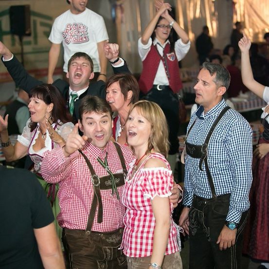 Eine Gruppe von Menschen in traditioneller deutscher Kleidung feiert auf einem Festival. Sie lächeln und scheinen sich zu amüsieren.