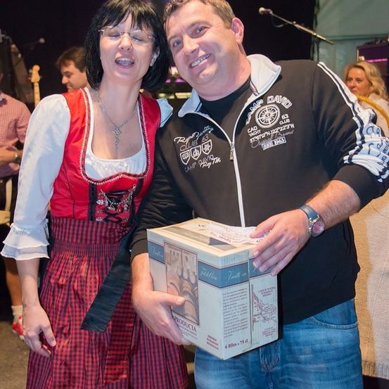 Ein Mann und eine Frau stehen bei einer Veranstaltung zusammen, der Mann hält eine Box und lächelt, während die Frau lacht und ein traditionelles Outfit trägt. Im Hintergrund sind verschwommen Menschen und eine Bühne mit einem Mikrofon zu sehen.
