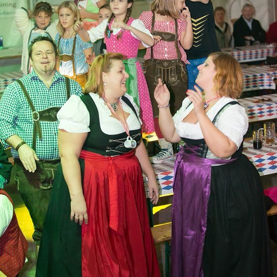 Eine Gruppe von Menschen in traditioneller bayerischer Kleidung versammelt sich in einer festlichen Umgebung, mit Kindern und Erwachsenen, die interagieren und lächeln. Der Hintergrund zeigt eine Party mit Tischen und Dekorationen.