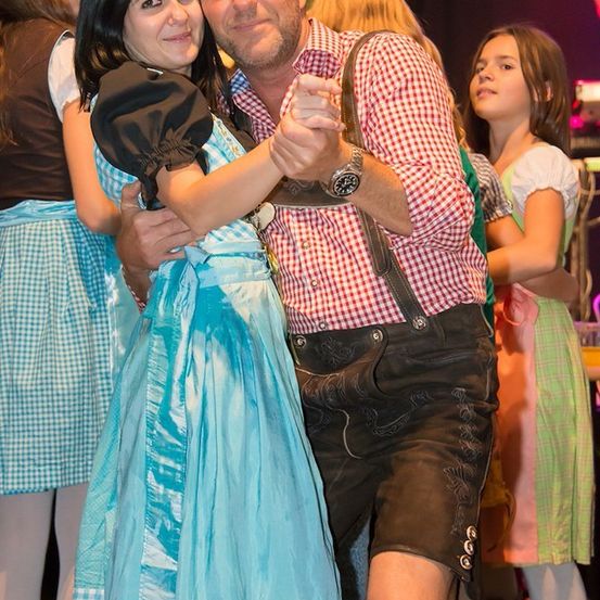 Ein Mann und eine Frau in traditioneller bayerischer Kleidung tanzen bei einem Fest, umgeben von anderen Festbesuchern.
