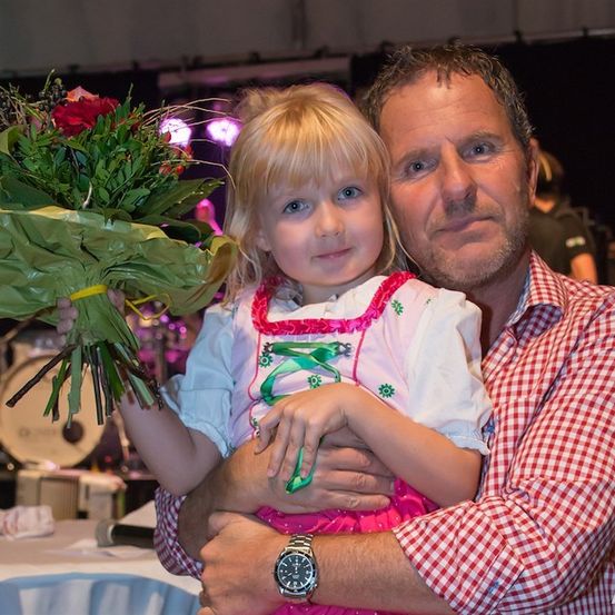 Ein Mann hält ein junges Mädchen in seinen Armen, beide lächeln für ein Foto. Das Mädchen hält einen Blumenstrauß. Dahinter befindet sich eine Bühne mit einem Schlagzeug und einer stehenden Person.
