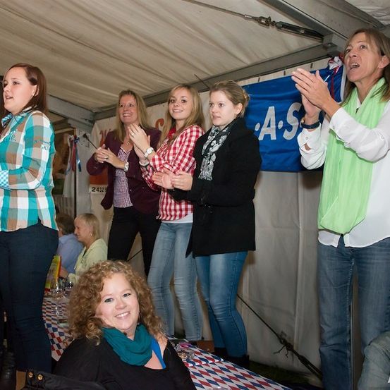 Eine Gruppe von Frauen in einem Zelt, einige klatschen, eine sitzt, mit einem Banner im Hintergrund, das 'A.S' steht.