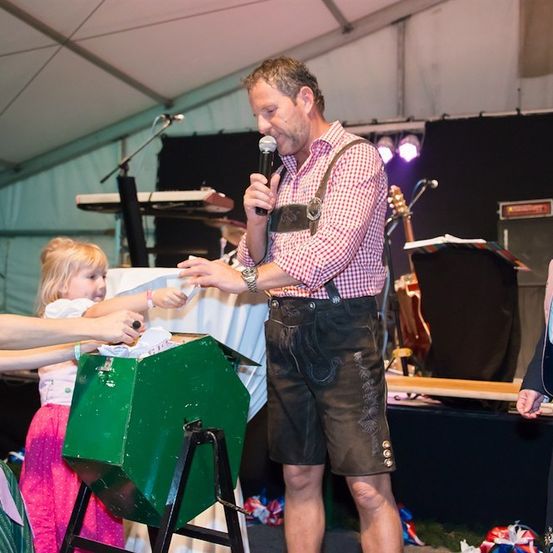 Ein Mann in traditioneller bayerischer Kleidung spricht in ein Mikrofon, während ein junges Mädchen ein Stück Papier in eine grüne Box legt. Dahinter ist eine Bühne mit Musikinstrumenten und einem schwarzen Hintergrund aufgebaut.