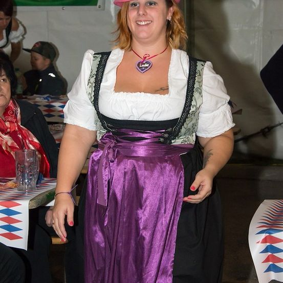 Eine Frau in traditioneller bayerischer Kleidung, einschließlich eines lila Rocks und eines rosa Huts, steht in einer festlichen Umgebung mit einem Banner im Hintergrund.