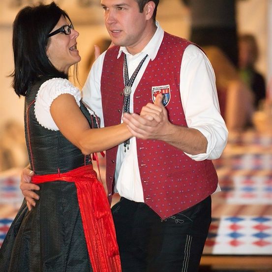 Ein Mann und eine Frau in traditioneller Kleidung tanzen bei einer Veranstaltung. Der Mann trägt eine rote Weste mit Logo, die Frau ein schwarzes Kleid mit rotem Gürtel. Sie lächeln und scheinen sich zu amüsieren.