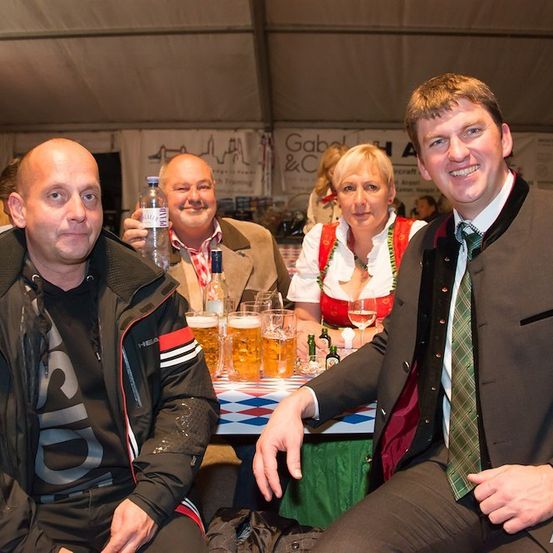 Vier Personen sitzen an einem Tisch mit Bierkrügen und Flaschen. Der Mann auf der rechten Seite trägt eine Krawatte und eine Uhr. Die Frau in der Mitte trägt ein traditionelles deutsches Kleid. Der Mann auf der linken Seite trägt eine Jacke. Der Mann in der Mitte hält eine Flasche.