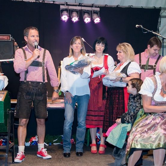 Eine Gruppe von Menschen in traditioneller deutscher Kleidung steht auf einer Bühne mit einem Mann, der ein Mikrofon hält. Hinter ihnen hält eine Frau einen Blumenstrauß.