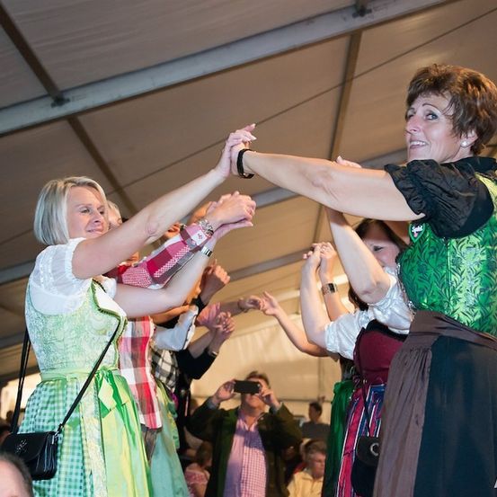 Eine Gruppe von Frauen in traditioneller Kleidung tanzt und klatscht in einem Zelt, während eine Menge zuschaut.