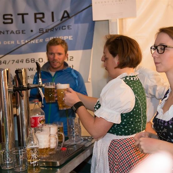 Zwei Frauen in traditioneller deutscher Kleidung zapfen Bier aus einem Zapfhahn bei einer Veranstaltung. Ein Mann steht hinter ihnen, und ein Banner mit Text ist im Hintergrund sichtbar.