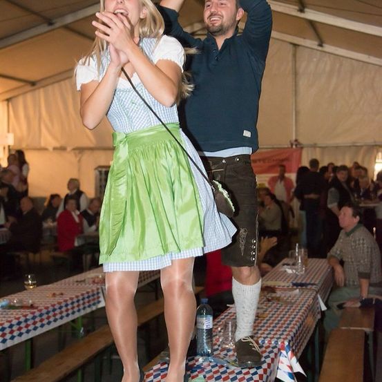 Eine Frau in einem grünen Kleid und ein Mann in traditioneller bayerischer Kleidung stehen auf einem Tisch, möglicherweise bei einem Fest auftretend oder feiernd.