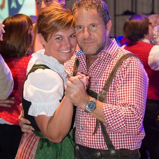 Ein Mann und eine Frau in traditioneller bayerischer Kleidung tanzen bei einer lebhaften Veranstaltung, umgeben von anderen Menschen in ähnlicher Kleidung.