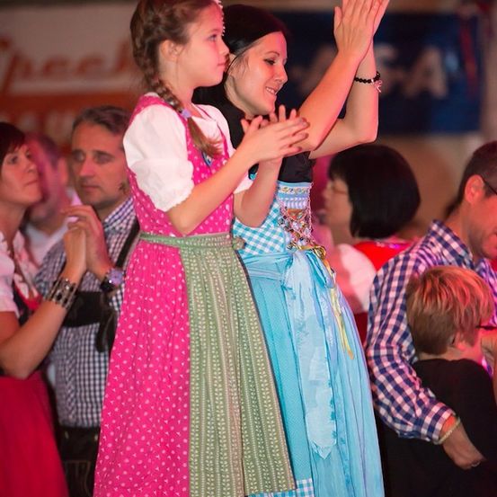 Zwei Mädchen in traditionellen bayerischen Kostümen tanzen und klatschen in einer festlichen Atmosphäre. Hinter ihnen schaut eine Menge Menschen zu, einige klatschen und andere lächeln. Im Hintergrund befindet sich ein Banner mit Text.