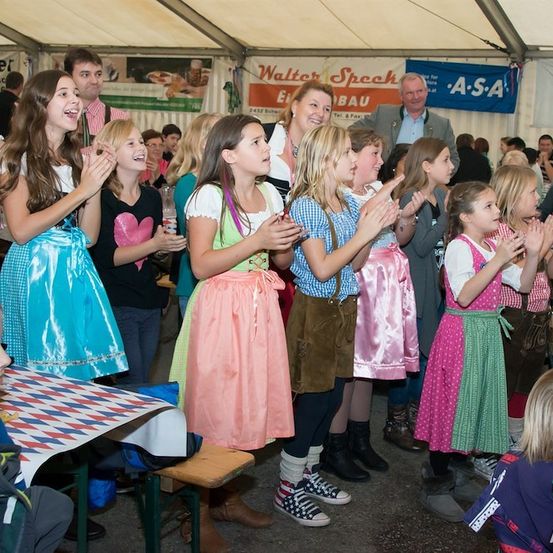 Eine Gruppe von Kindern und Erwachsenen klatscht und lächelt bei einer Veranstaltung im Freien, möglicherweise einem Festival. Einige tragen traditionelle Kleidung. Im Hintergrund befinden sich Zelte und Banner.