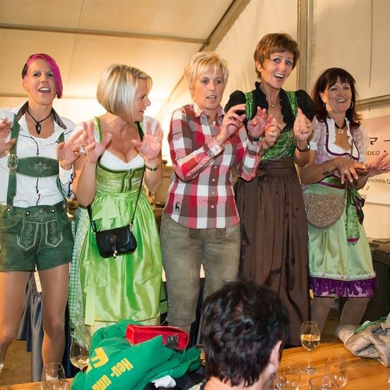 Fünf Frauen in traditioneller deutscher Kleidung, wahrscheinlich auf einem Festival, lächeln und machen Handgesten. Sie stehen vor einem Tisch mit Weingläsern und einem Banner im Hintergrund.