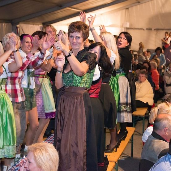 Eine Gruppe von Frauen in traditioneller deutscher Kleidung klatscht und jubelt in einem überfüllten Raum.