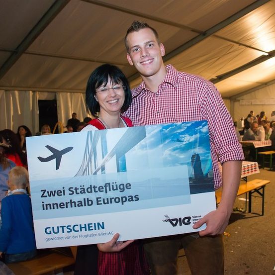 Ein Mann und eine Frau halten ein Zertifikat für zwei Städteflüge innerhalb Europas. Sie lächeln und stehen in einem Zelt mit einer Menschenmenge im Hintergrund.