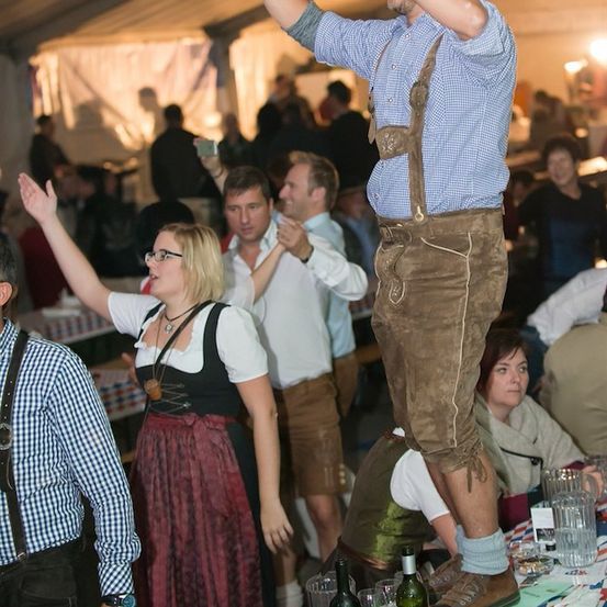 Eine Gruppe von Menschen in traditioneller bayerischer Kleidung tanzt und feiert in einem Zelt. Ein Mann steht auf einem Tisch und hebt die Hände, während andere klatschen und jubeln.