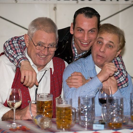 Drei Männer sitzen an einem Tisch mit verschiedenen Getränken, einer der Männer hat seinen Arm um die Schultern der anderen.