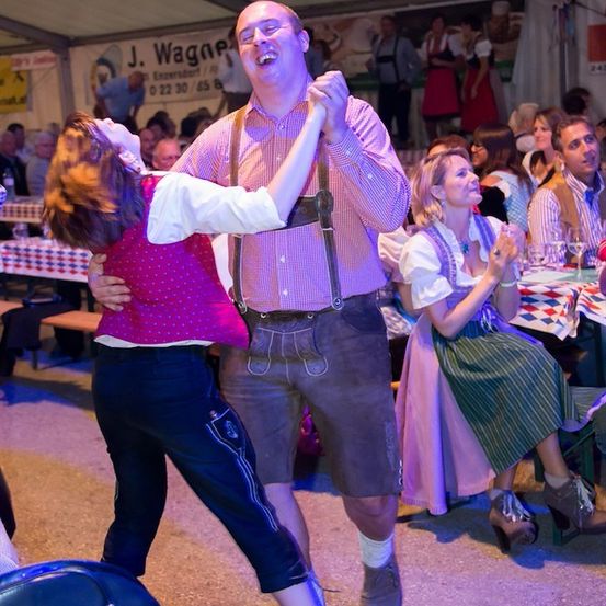 Ein Mann und eine Frau tanzen in traditioneller bayerischer Kleidung auf einem lebhaften Festival, umgeben von anderen Feiernden, die die Atmosphäre genießen.