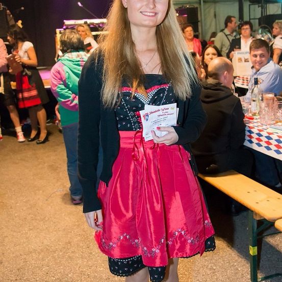 Eine Frau in einem rosa Kleid mit einem schwarzen Oberteil und einer weißen Karte in der Hand steht in einem überfüllten Raum.