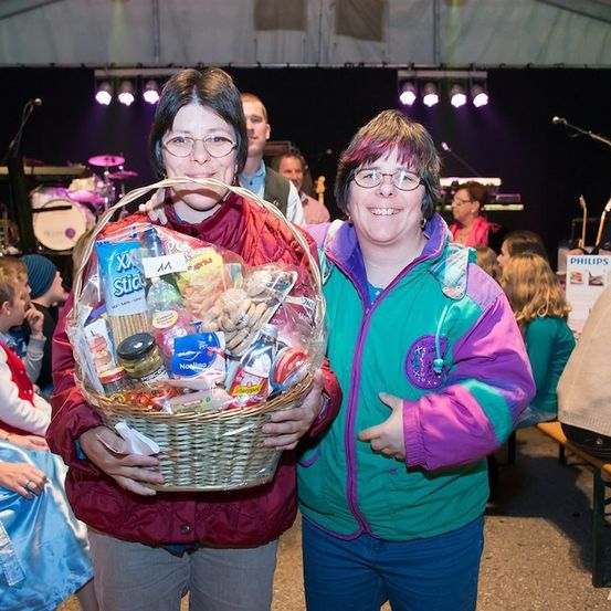 Zwei Frauen stehen zusammen und halten einen Korb mit Snacks und Getränken bei einer Veranstaltung. Dahinter spielt eine Band auf der Bühne.
