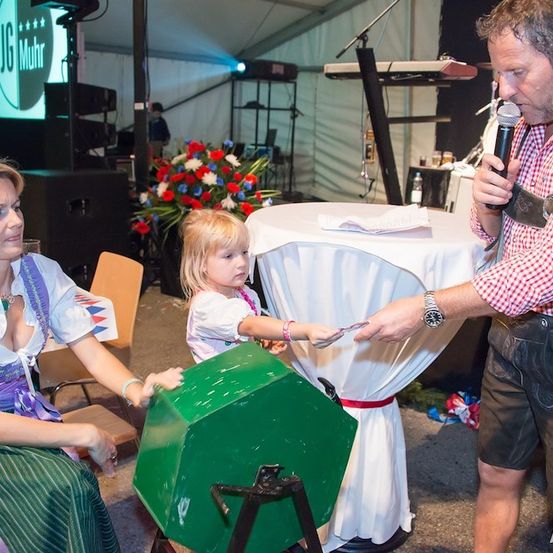 Ein Mann und eine Frau in traditioneller bayerischer Kleidung stehen neben einem grünen Objekt, während ein kleines Mädchen einen Schlüssel hält. Der Mann hält ein Mikrofon und scheint zu sprechen. Hinter ihnen ist ein Tisch mit weißer Tischdecke und einer Blumenarrangement zu sehen.