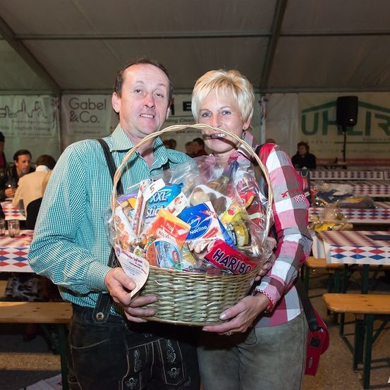 Ein Mann und eine Frau stehen zusammen und halten einen Korb voller verschiedener Snacks, darunter Haribo und andere Marken, in einer Indoor-Veranstaltung mit Tischen und Menschen im Hintergrund.