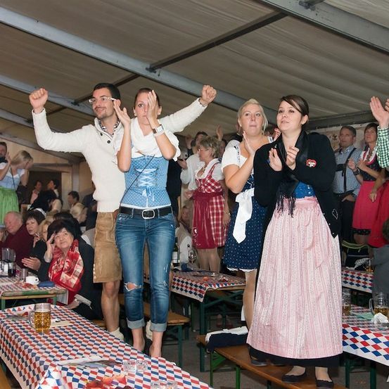 Eine Gruppe von Menschen in traditioneller bayerischer Kleidung klatscht und jubelt in einem großen Zelt, möglicherweise bei einem Festival. Tische sind mit Bierkrügen und karierten Tischdecken ausgestattet.