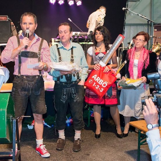 Eine Gruppe von Menschen in traditioneller bayerischer Kleidung steht auf einer Bühne, einer hält ein Mikrofon und ein anderer eine rote Tasche mit der Aufschrift Arbo. Dahinter macht eine Person ein Foto.