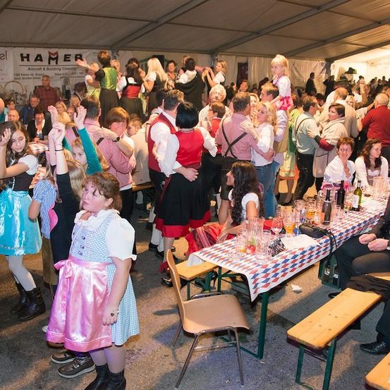 Eine große Gruppe von Menschen in traditioneller bayerischer Kleidung versammelt sich in einem Zelt, einige tanzen und andere sitzen an Tischen mit Getränken und Essen. Ein Banner mit dem Text 'HAMER & Co.' ist sichtbar.