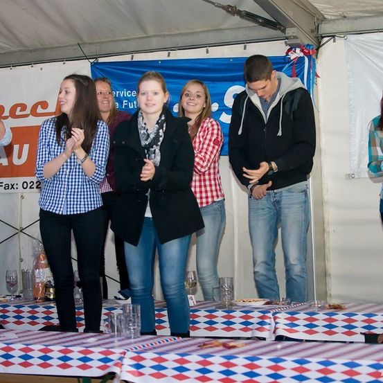 Eine Gruppe von Menschen steht auf einer Bühne unter einem Zelt, klatscht und lächelt. Sie sind lässig gekleidet und scheinen sich auf einer Veranstaltung zu befinden. Vor ihnen stehen Tische mit rot-weiß-blauen Tischdecken.