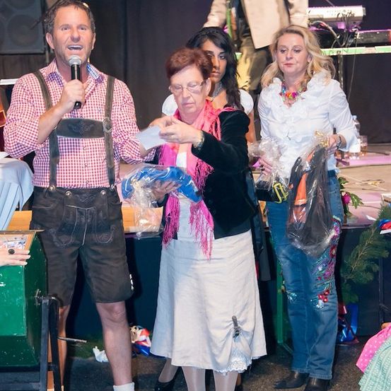 Ein Mann in traditioneller bayerischer Kleidung überreicht einer Frau ein Geschenk, während eine Menge zusieht. Dahinter hält eine Frau ein eingewickeltes Geschenk und ein Mann steht neben einer Tastatur.
