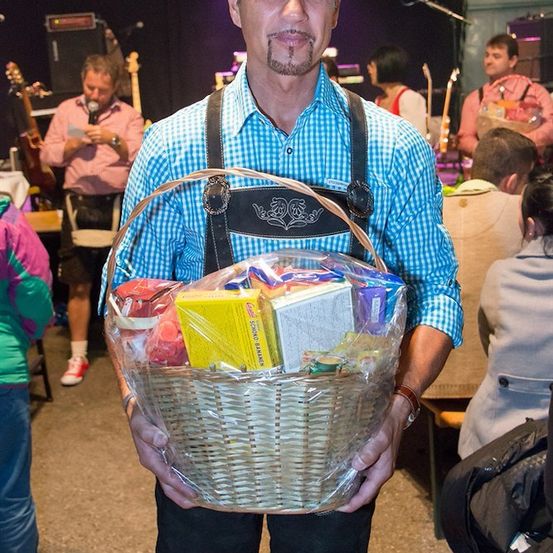 Ein Mann in traditioneller Kleidung hält einen Korb mit verschiedenen Gegenständen und steht in einem Raum mit einer Band im Hintergrund.