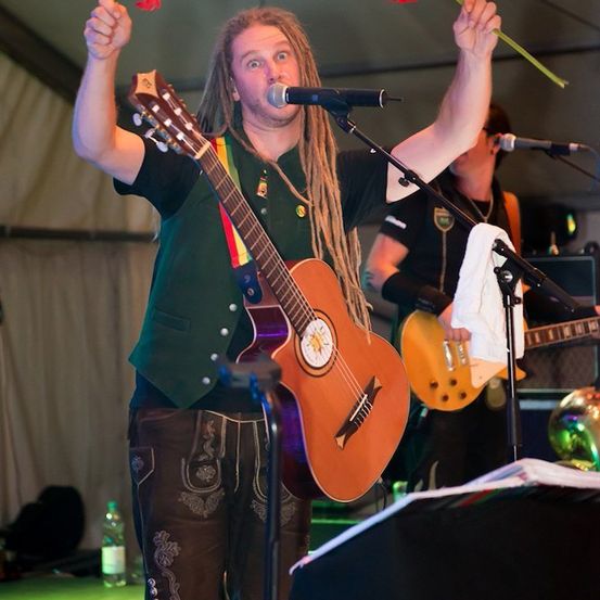 Ein Mann mit Dreadlocks tritt auf der Bühne auf, hält eine Gitarre und eine rote Blume in der Hand, während ein anderer Musiker Gitarre spielt.