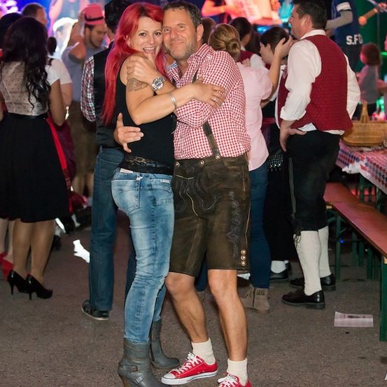 Ein Mann und eine Frau tanzen eng beieinander auf einer lebhaften Veranstaltung im Freien, umgeben von anderen Menschen in traditioneller Kleidung.