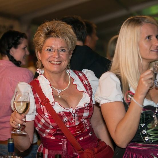 Zwei Frauen in traditioneller bayerischer Kleidung, eine mit einem Weinglas und die andere mit einem Bierkrug, lächeln und schauen bei einer festlichen Veranstaltung nach vorne.
