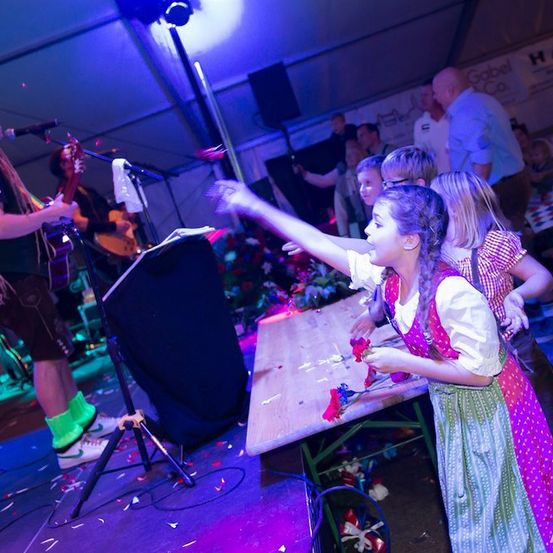 Ein junges Mädchen in einem traditionellen Kleid wirft Blumen auf die Bühne, während eine Band im Hintergrund bei einem festlichen Ereignis spielt.