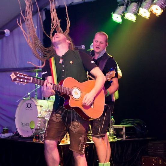 Ein Mann mit Dreadlocks spielt Gitarre auf der Bühne, während ein anderer Mann hinter ihm steht. Die Bühne ist mit grünen und weißen Lichtern beleuchtet, und es gibt ein Schlagzeug und Flaschen auf der Bühne.