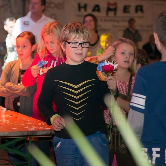 Eine Gruppe von Kindern, darunter ein Junge mit Brille, der eine Blume hält, steht in einem Raum mit Erwachsenen und einem Banner mit der Aufschrift 'HAMER'.